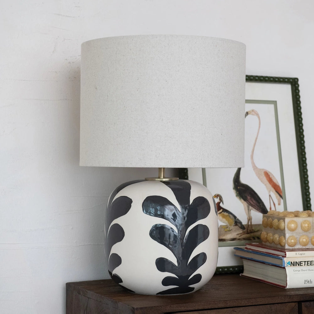 Stoneware table lamp with a black abstract leaf design and white linen shade on a wooden dresser.