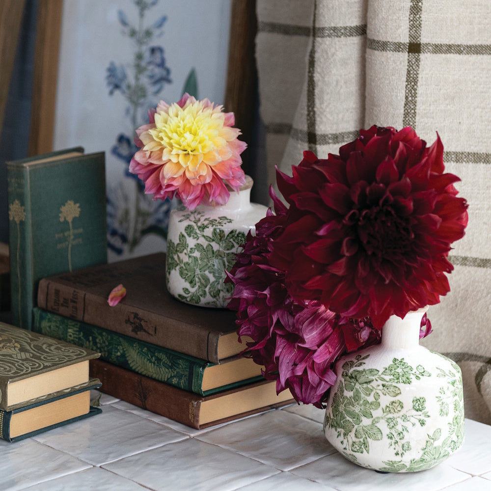 Two patterned vases on a tiled surface holding vibrant dahlias next to a stack of vintage books.