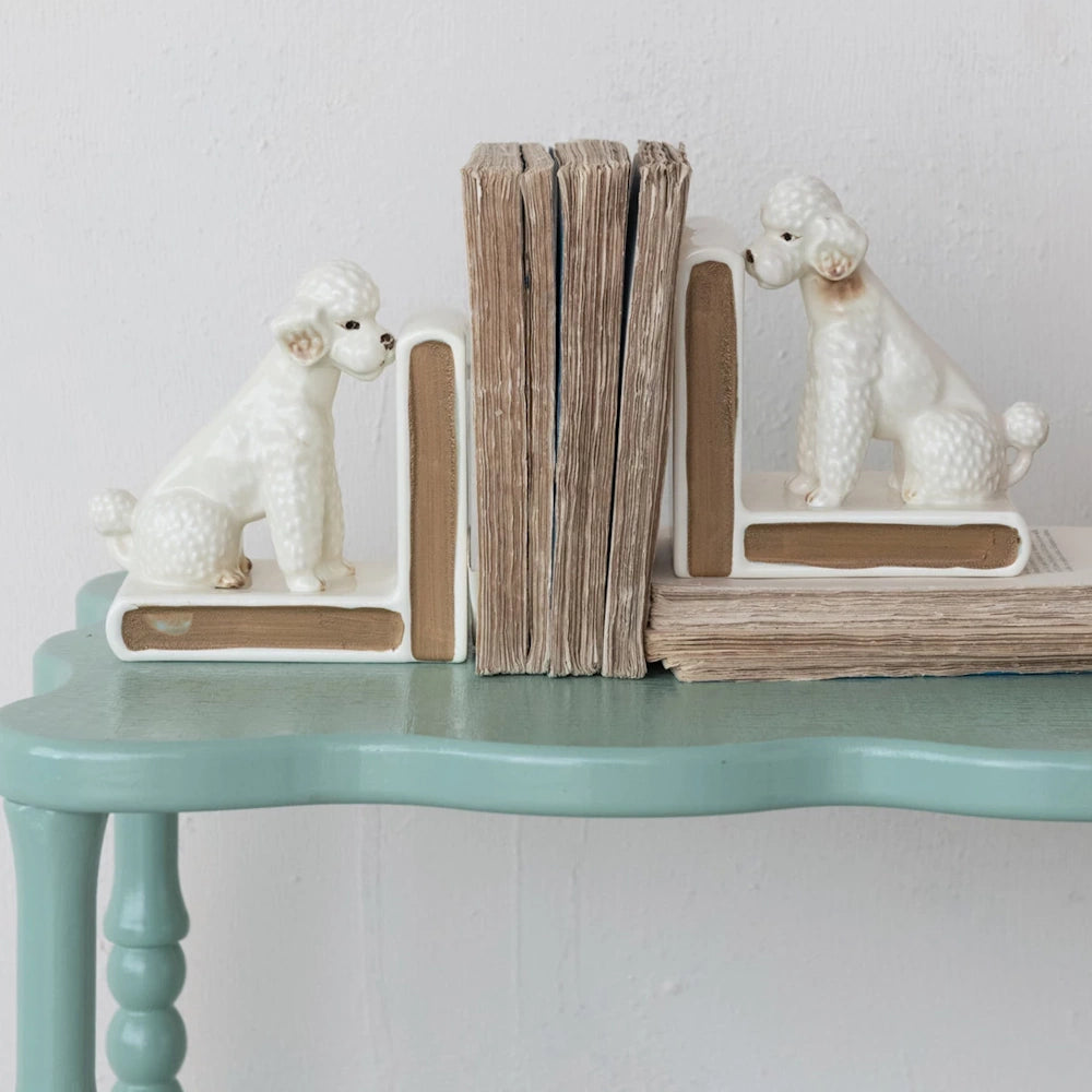 Vintage white ceramic poodle bookends holding old books on a light blue scalloped table.