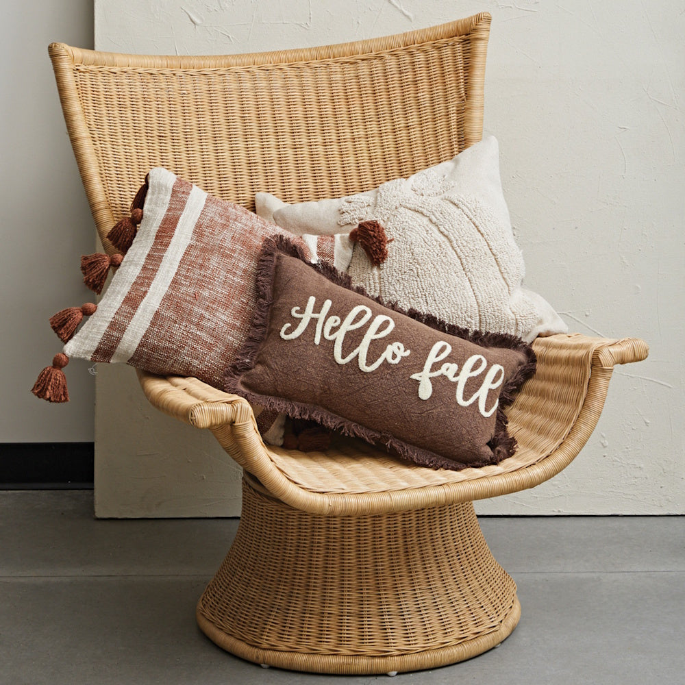 Three autumn pillows, including a brown 'Hello fall' cushion, arranged on a woven wicker chair.
