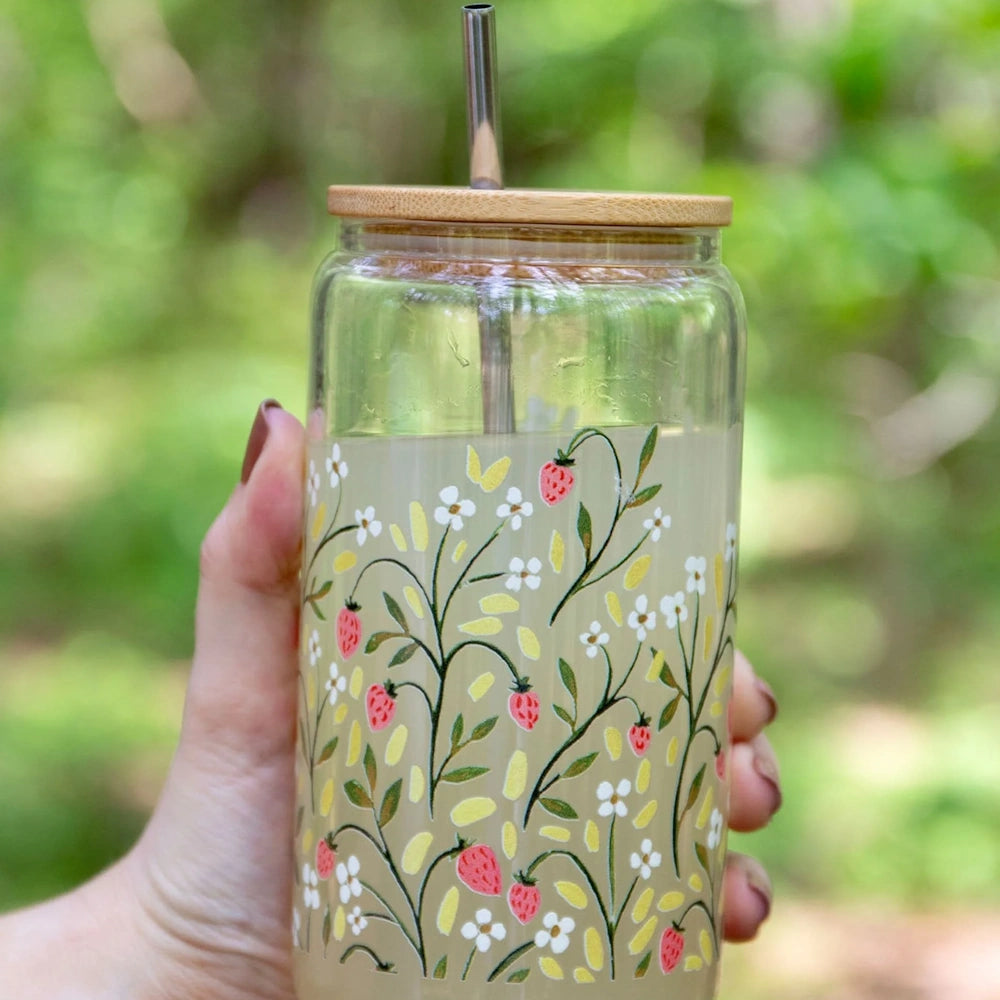 A hand holding a glass tumbler with strawberry and flower illustrations, a bamboo lid, and a metal straw.
