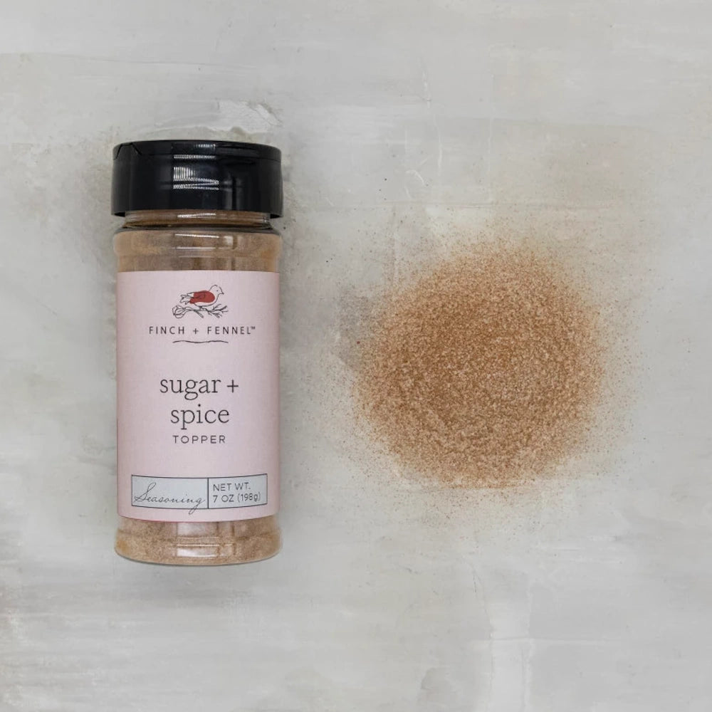 Bottle of Finch + Fennel Sugar + Spice Topper next to a pile of the fine brown seasoning on a light marbled surface.