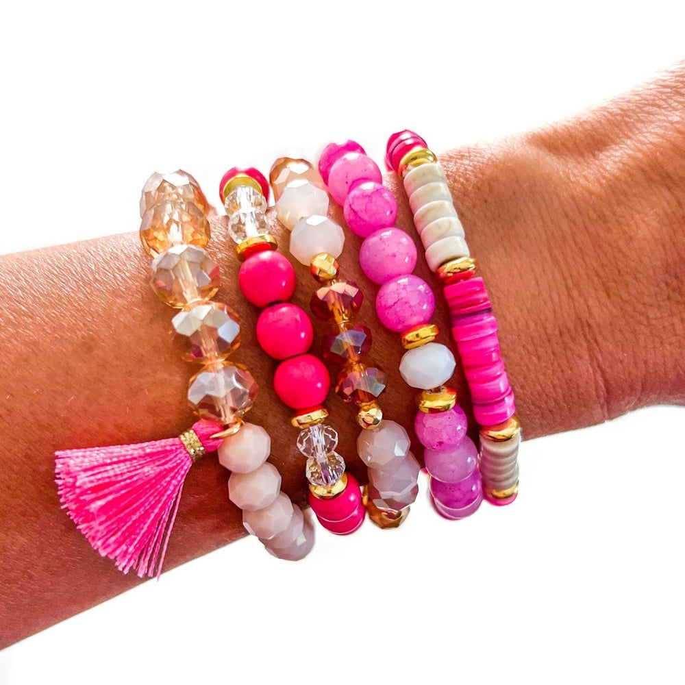 A stack of vibrant pink, white, and gold beaded bracelets worn on a wrist, featuring a pink tassel.