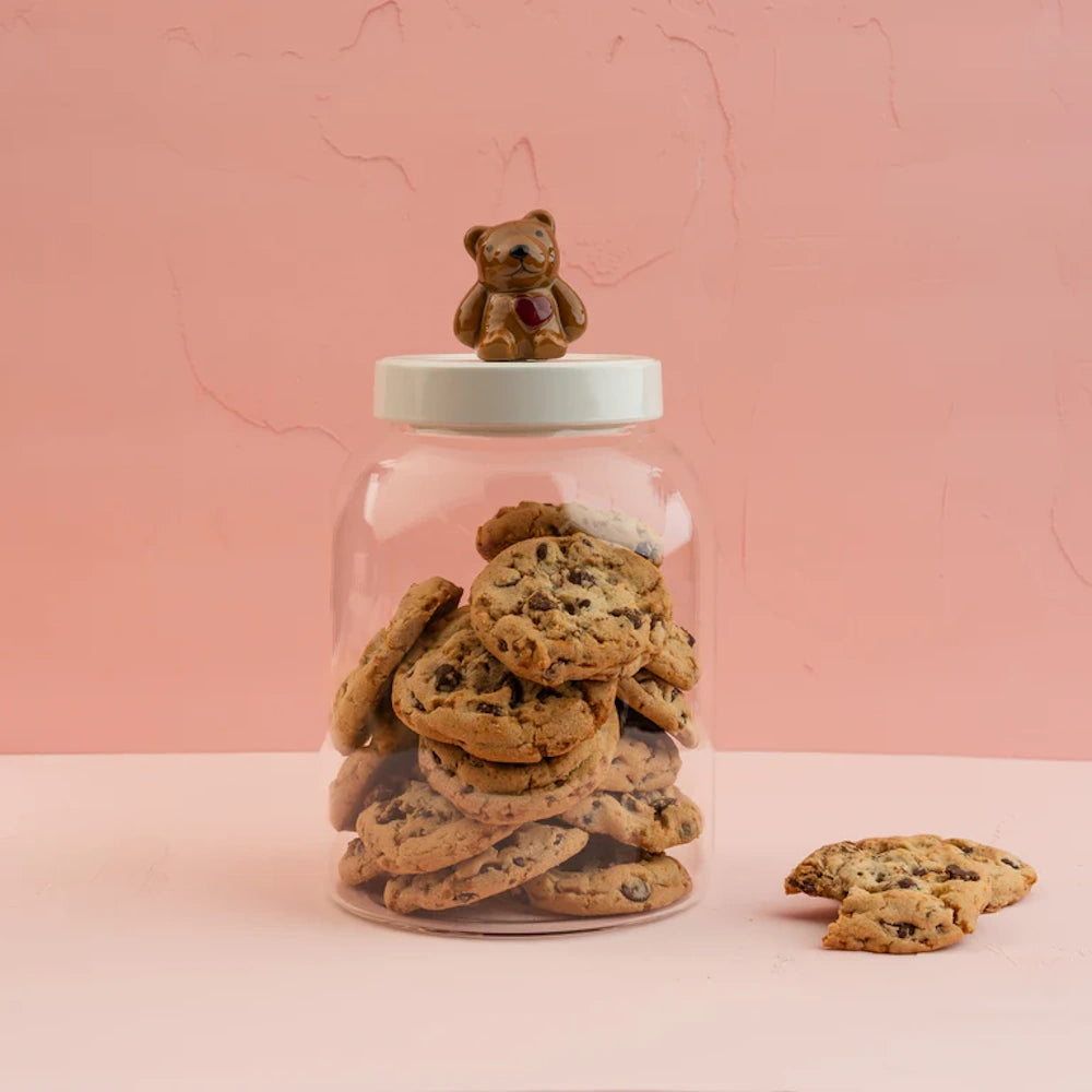 Glass cookie jar filled with chocolate chip cookies topped with a ceramic bear figurine against a pink background.