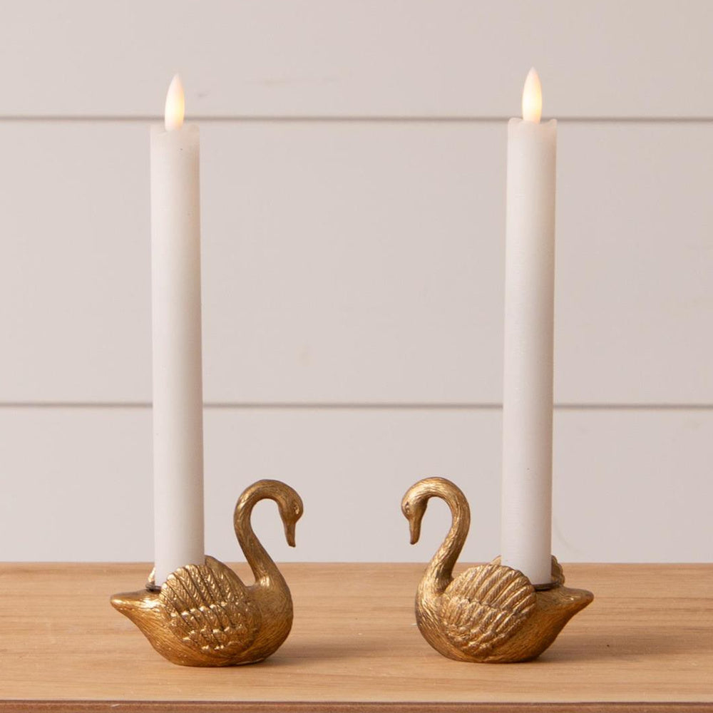 Pair of gold swan tapered candle holders with white candles on a wooden surface, elegant home decor accent.