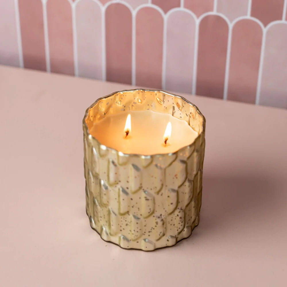 A double-wick candle burning in a textured gold glass jar on a pink surface against a scalloped tile background.