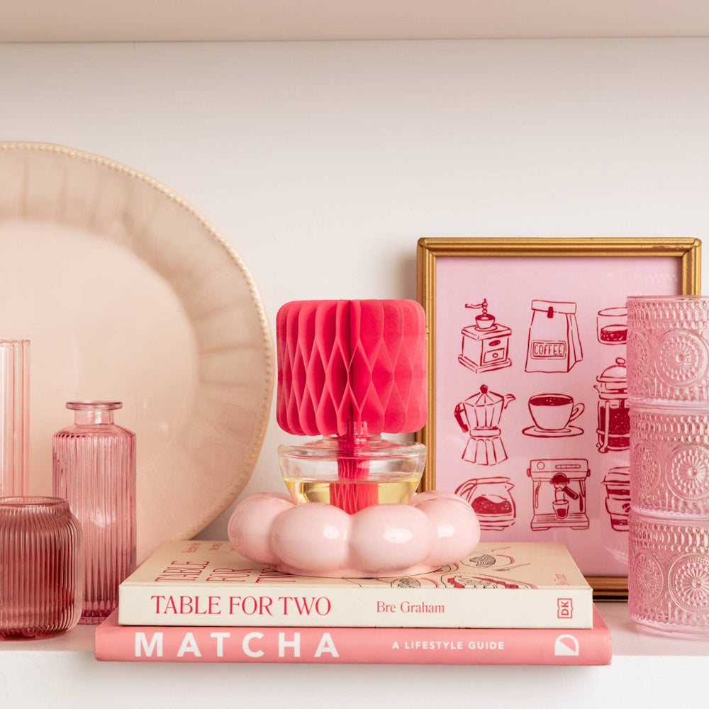 Pink honeycomb fragrance diffuser on books alongside pink glass decor and framed art on a shelf.