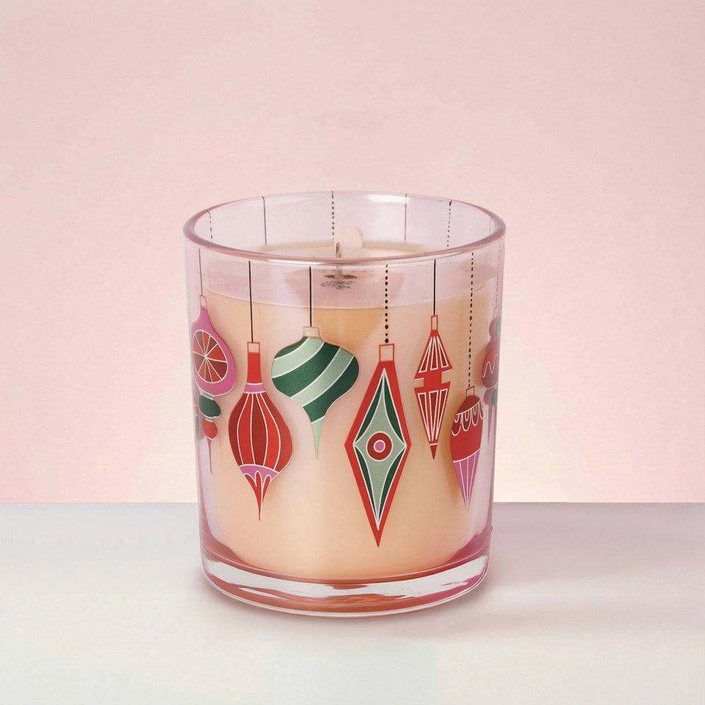 Sweet Grace soy candle in a glass jar decorated with colorful holiday ornaments on a light pink background.
