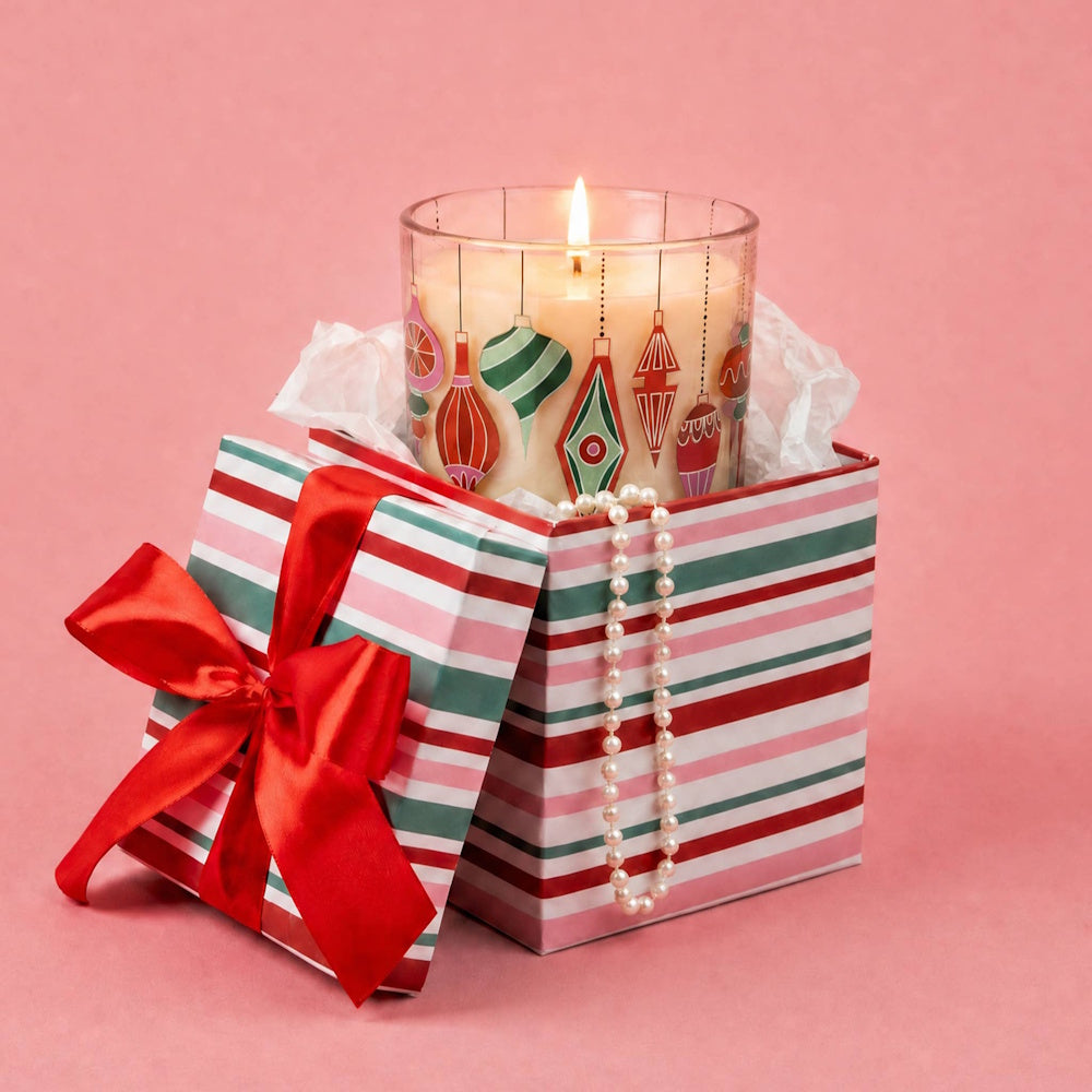 Lit candle with ornament design inside a striped gift box with a red bow and pearl necklace against pink.