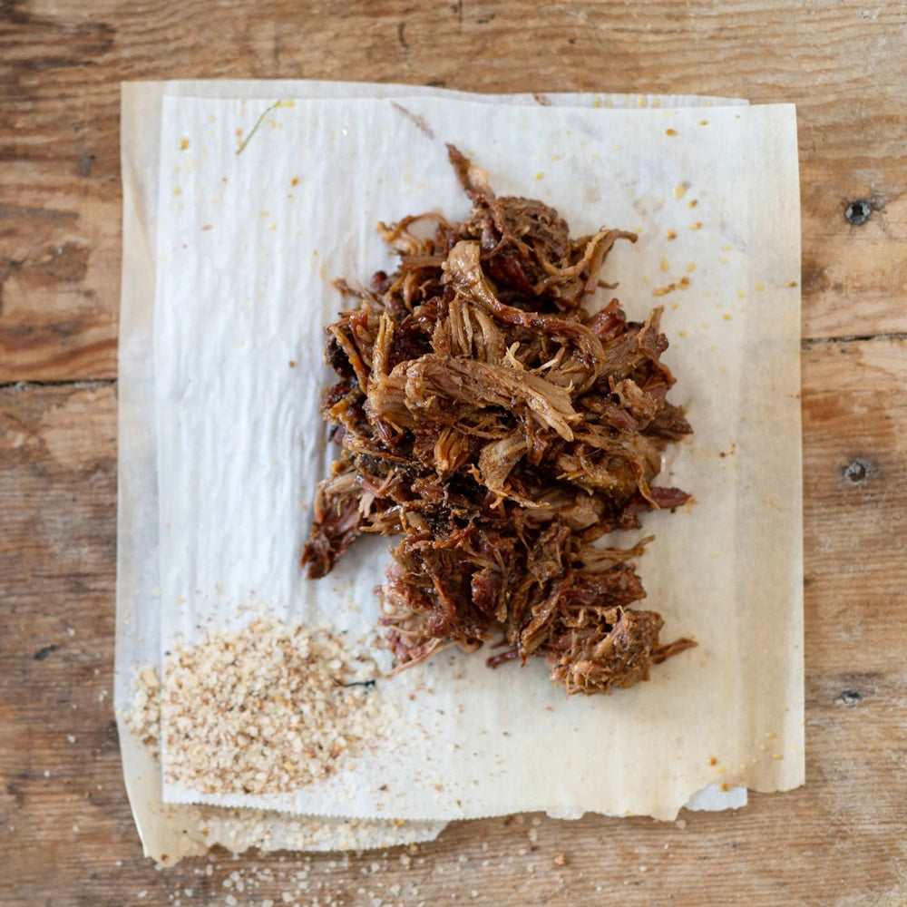 Pulled pork pile on parchment paper over a rustic wood surface with a side of dry rub seasoning.