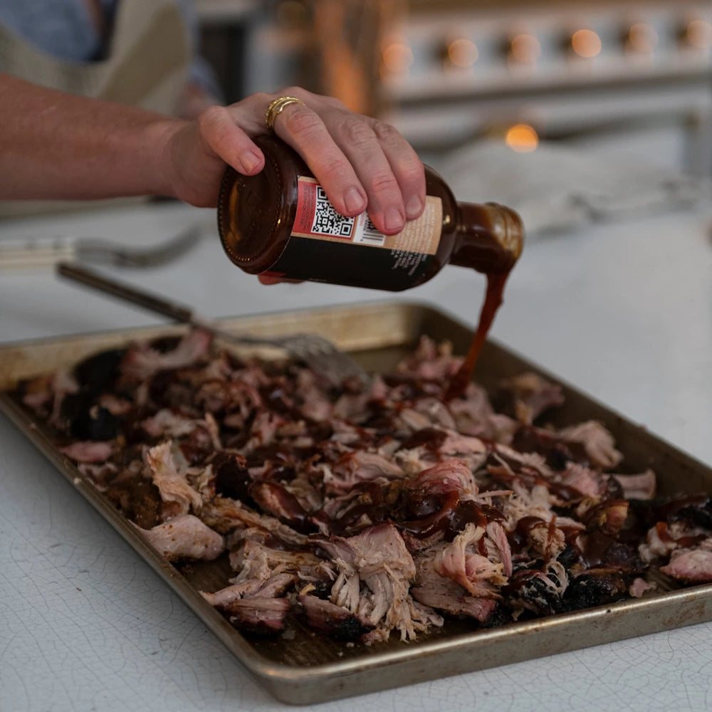 A hand pouring dark honey habanero BBQ sauce from a bottle onto a tray of shredded pulled pork in a kitchen.