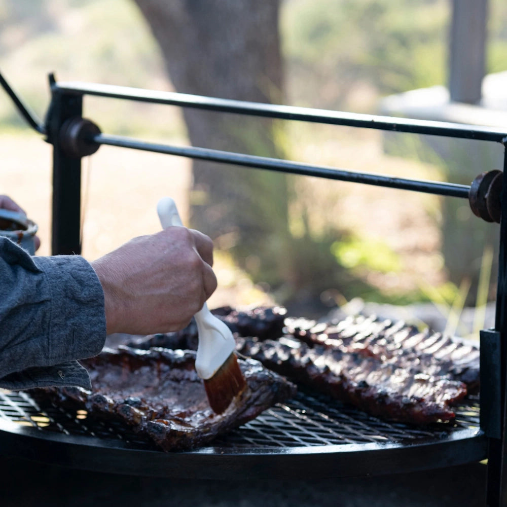 A person brushes honey habanero BBQ sauce onto racks of ribs grilling on an outdoor charcoal grill.