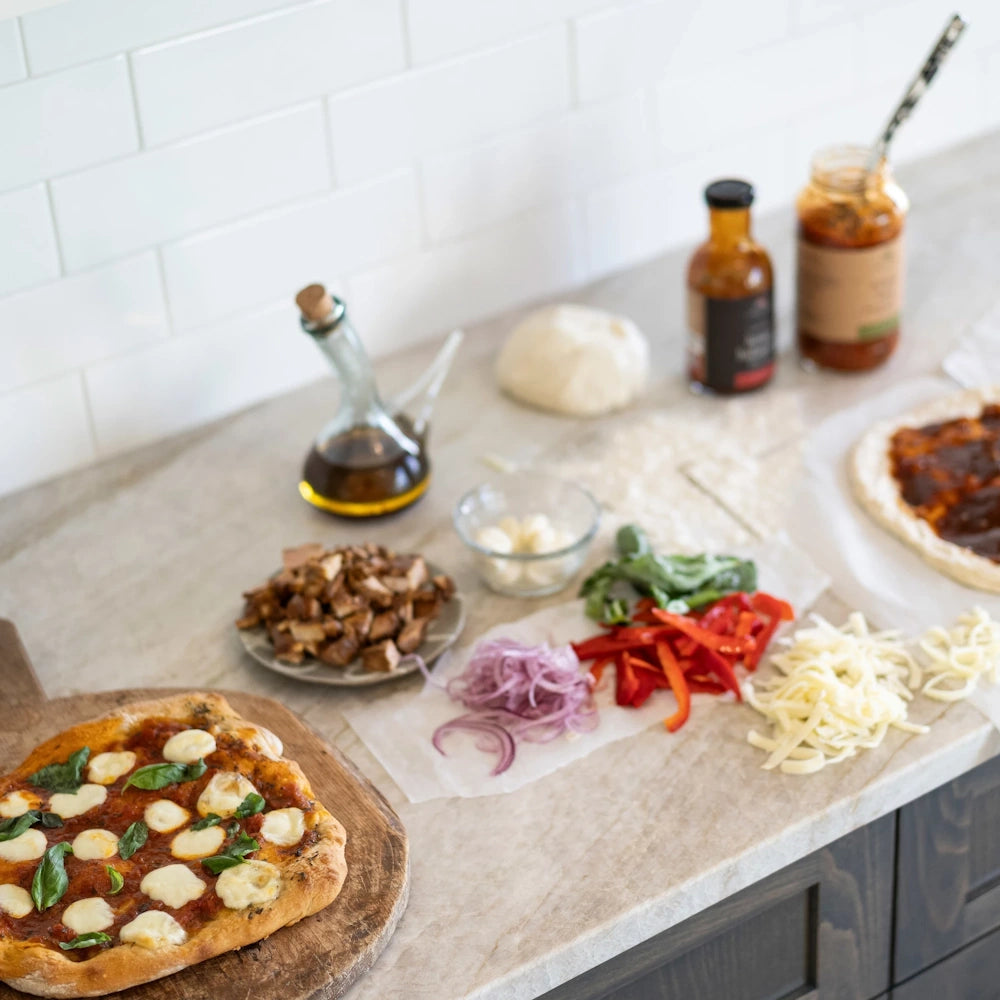 Homemade pizza with fresh mozzarella, basil, and red peppers on a marble counter with prep ingredients.