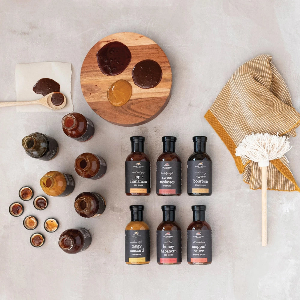 Assorted Finch & Fennel BBQ sauce bottles with sauce samples on a wooden board, mop brush, and striped towel.