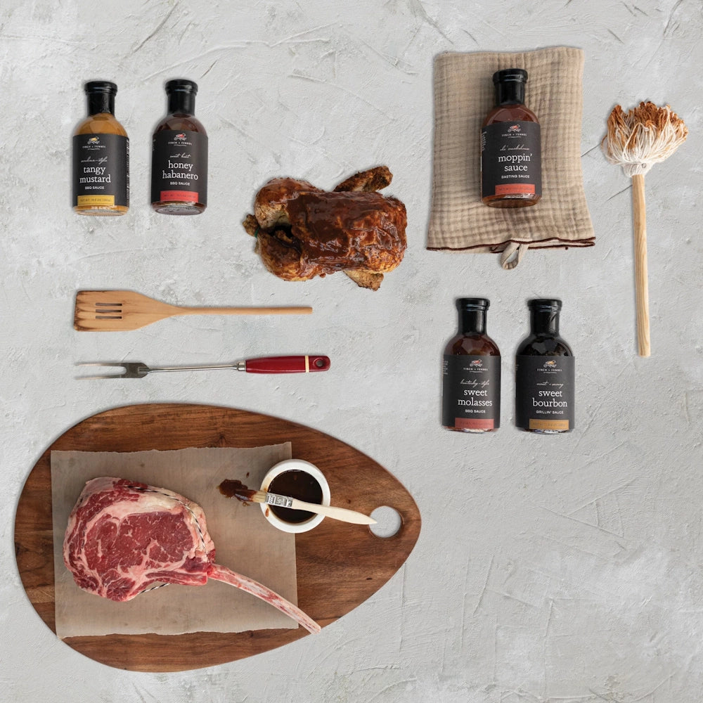 Flat lay of BBQ sauces, a tomahawk steak, roasted chicken, and grilling tools on a light textured surface.