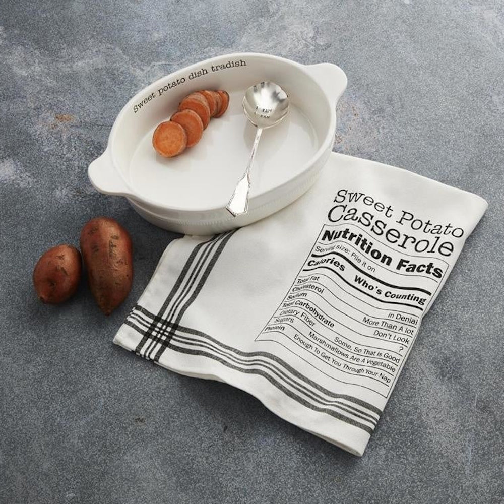 White oval ceramic baking dish with sliced sweet potatoes, serving spoon, and nutrition facts towel.