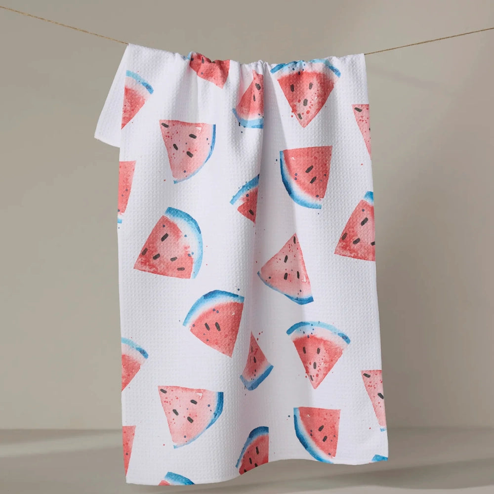 A white waffle-weave kitchen towel patterned with watercolor watermelon slices hangs on a clothesline.