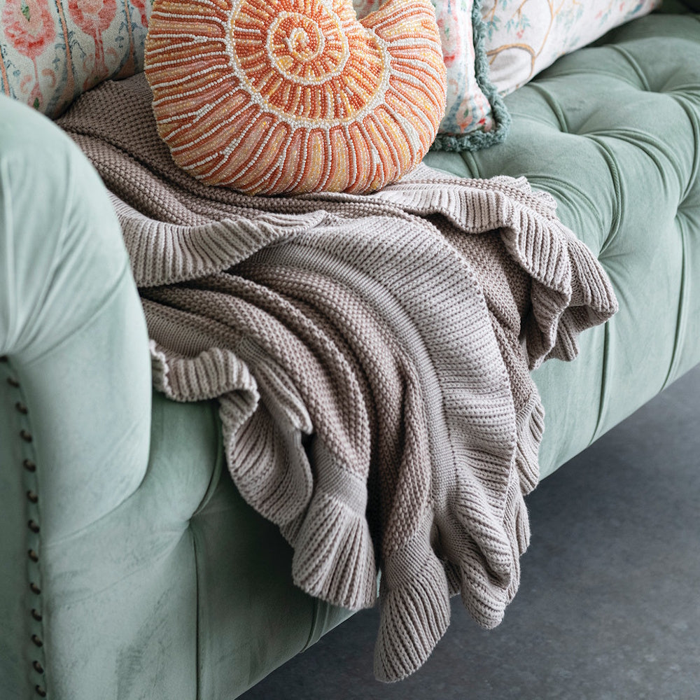 Taupe cotton knit throw blanket with ruffle edge draped on a sage green tufted sofa.