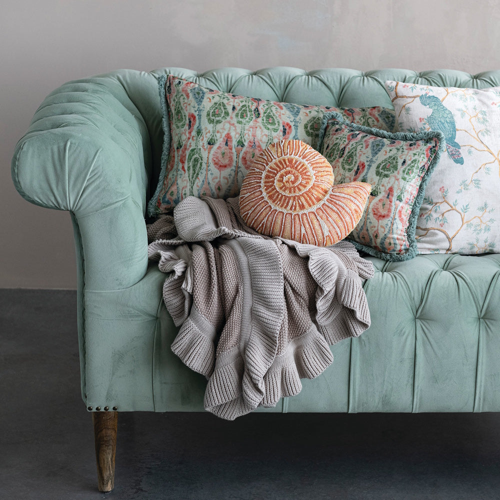 Taupe cotton knit throw blanket with ruffle trim styled on a mint green tufted sofa.
