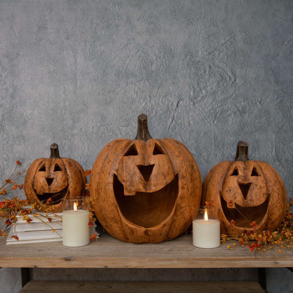 Three carved terracotta jack-o'-lanterns with wide mouths sit on a wooden shelf beside lit glass candles.