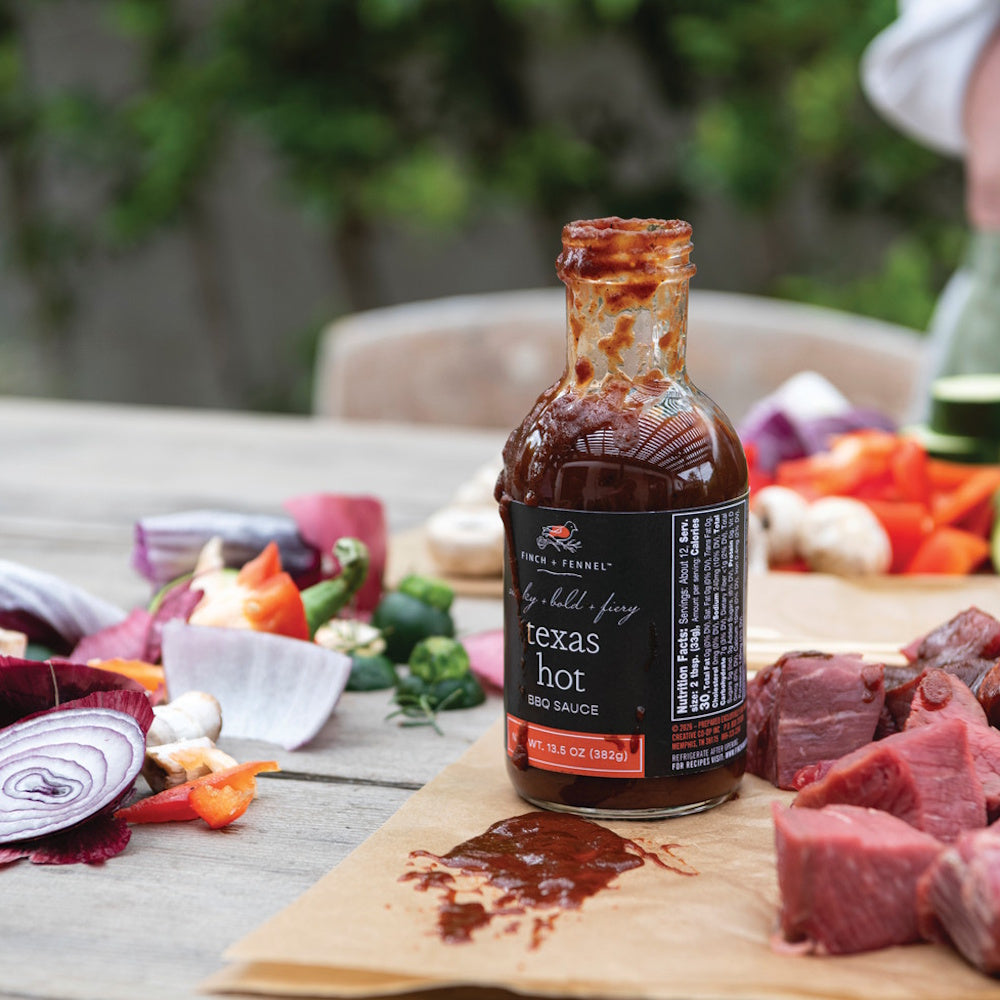 Finch + Fennel Texas Hot BBQ Sauce bottle on a table with raw cubed meat and chopped vegetables.