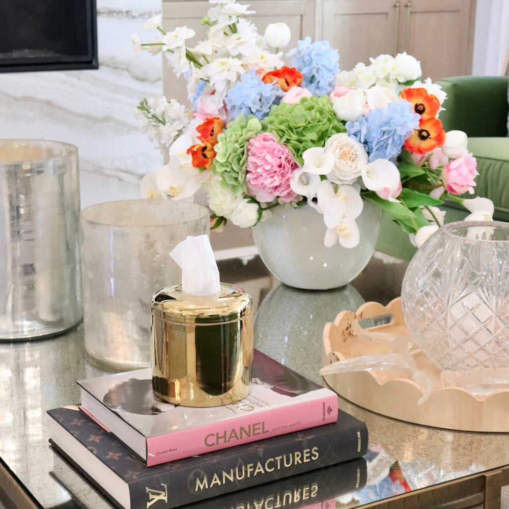 Gold tissue dispenser on stacked books surrounded by glass cylinders and a colorful floral arrangement.
