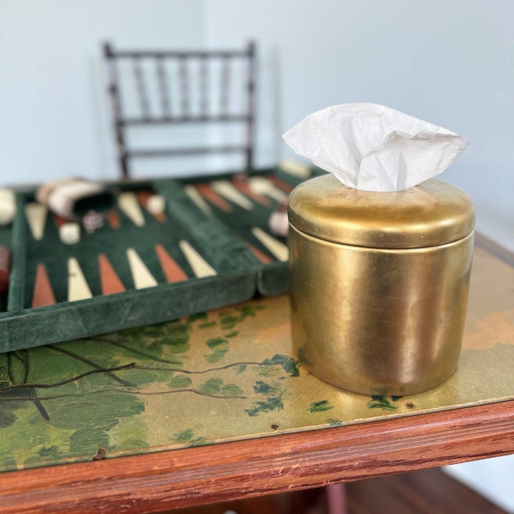 Antique gold cylindrical tissue box on a wooden table with a backgammon set and a chair in the background.
