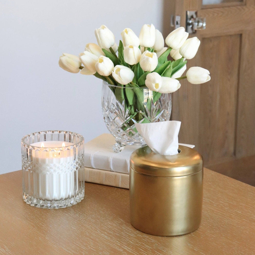 A gold cylindrical tissue dispenser, a crystal vase of white tulips, and a lit candle on a wooden table.