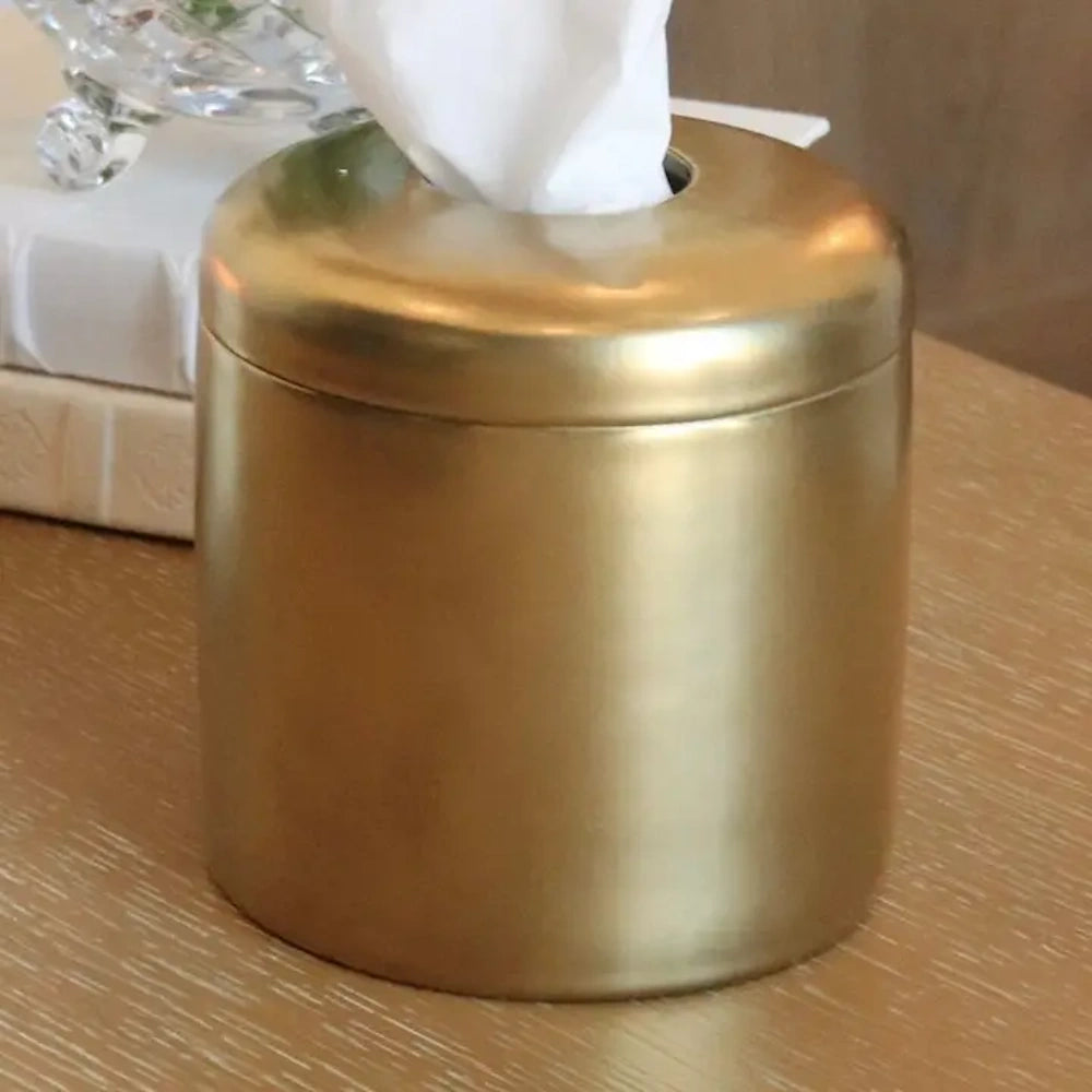 A cylindrical brushed gold tissue box cover with a domed lid sitting on a light wood surface.