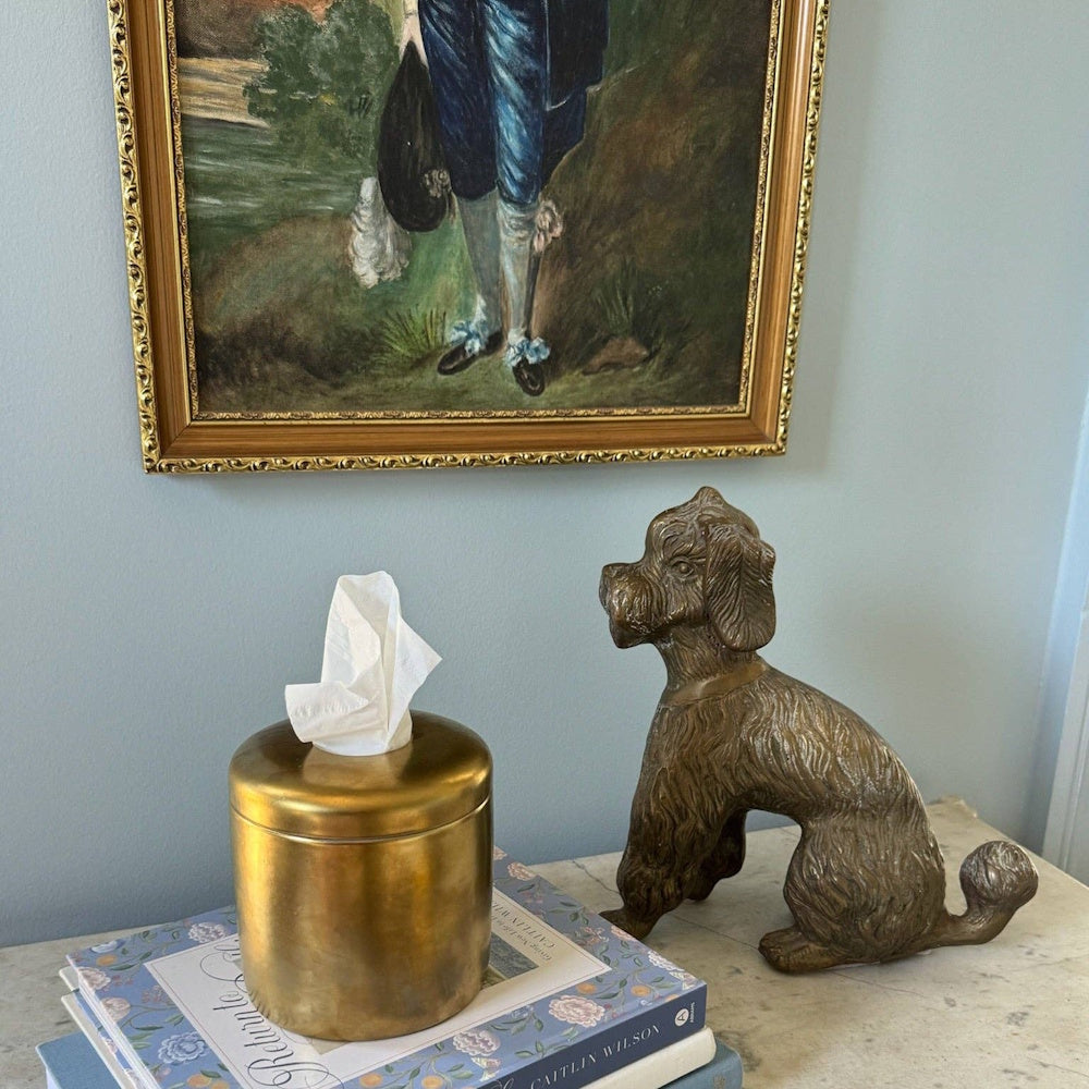 A gold cylindrical tissue box sits on a stack of books next to a bronze dog statue beneath a framed painting.