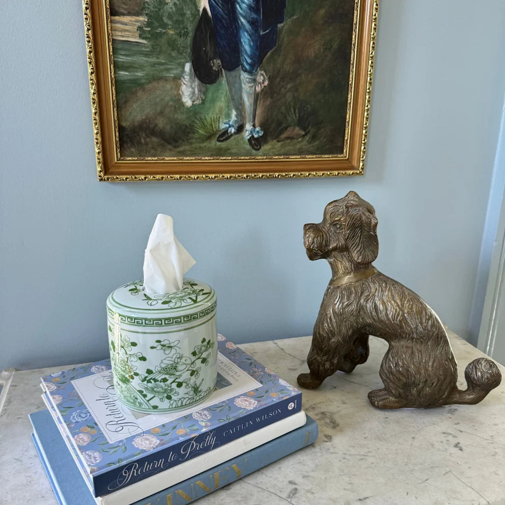Green floral chinoiserie tissue box on books next to a brass dog statue and framed art in a classic interior setting.