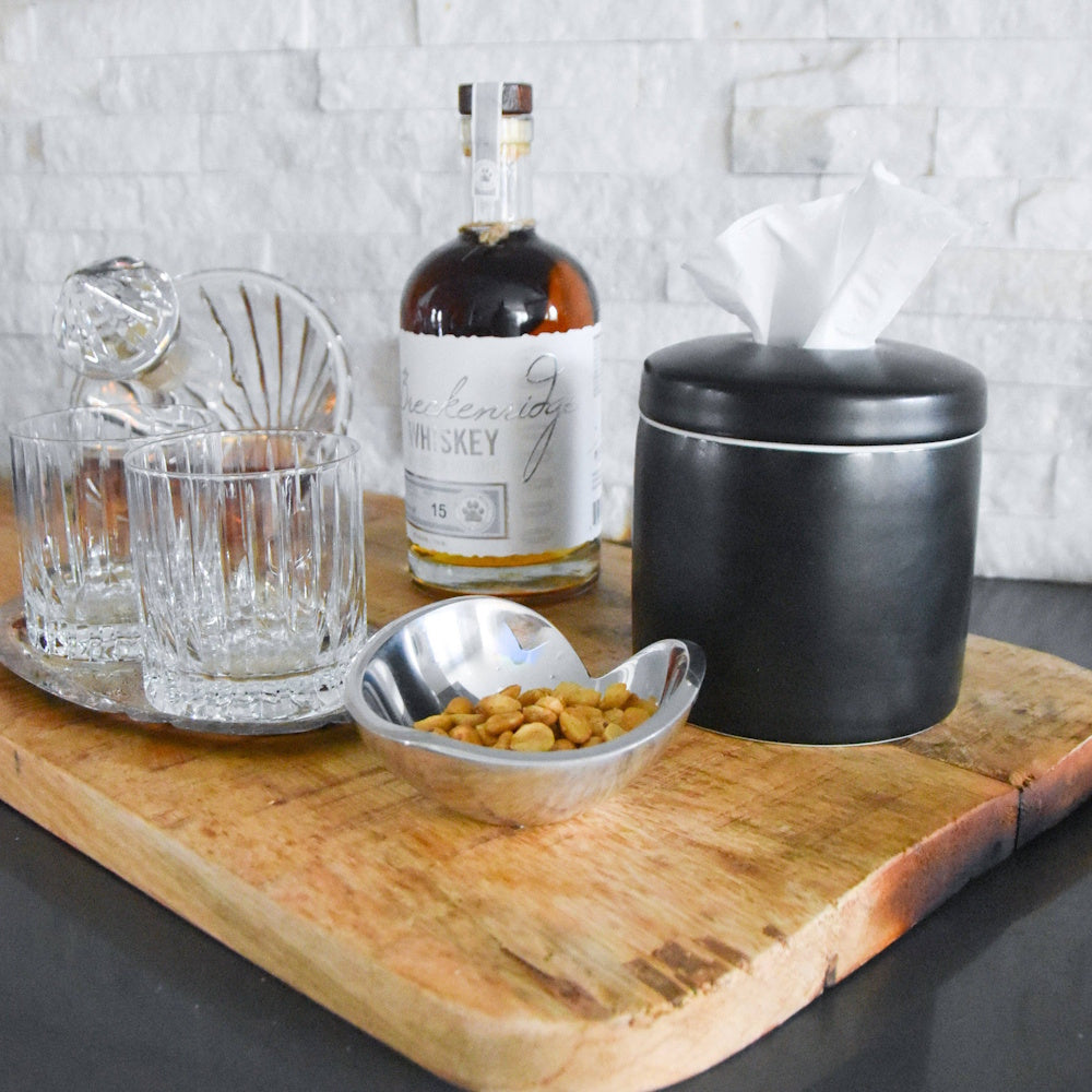 Matte black tissue dispenser on a wooden board next to a whiskey bottle, glasses, and a small bowl of nuts.