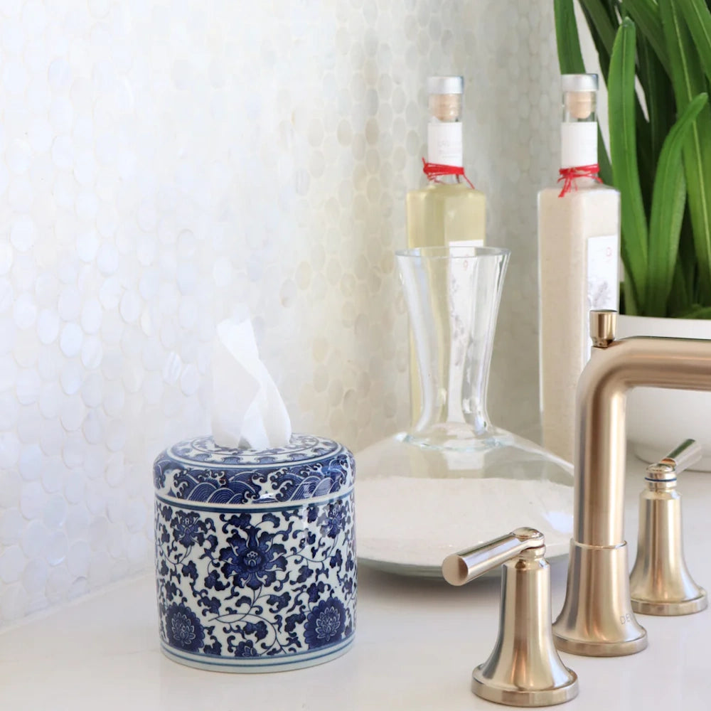 Blue and white floral ceramic tissue vessel on a bathroom counter with a gold faucet and pearl tile backsplash.