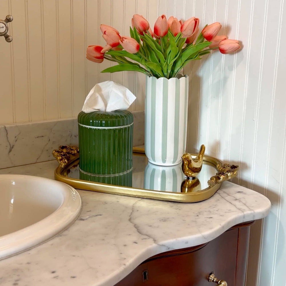 Green ribbed tissue dispenser on a mirrored tray with a striped vase of tulips and a small gold bird.