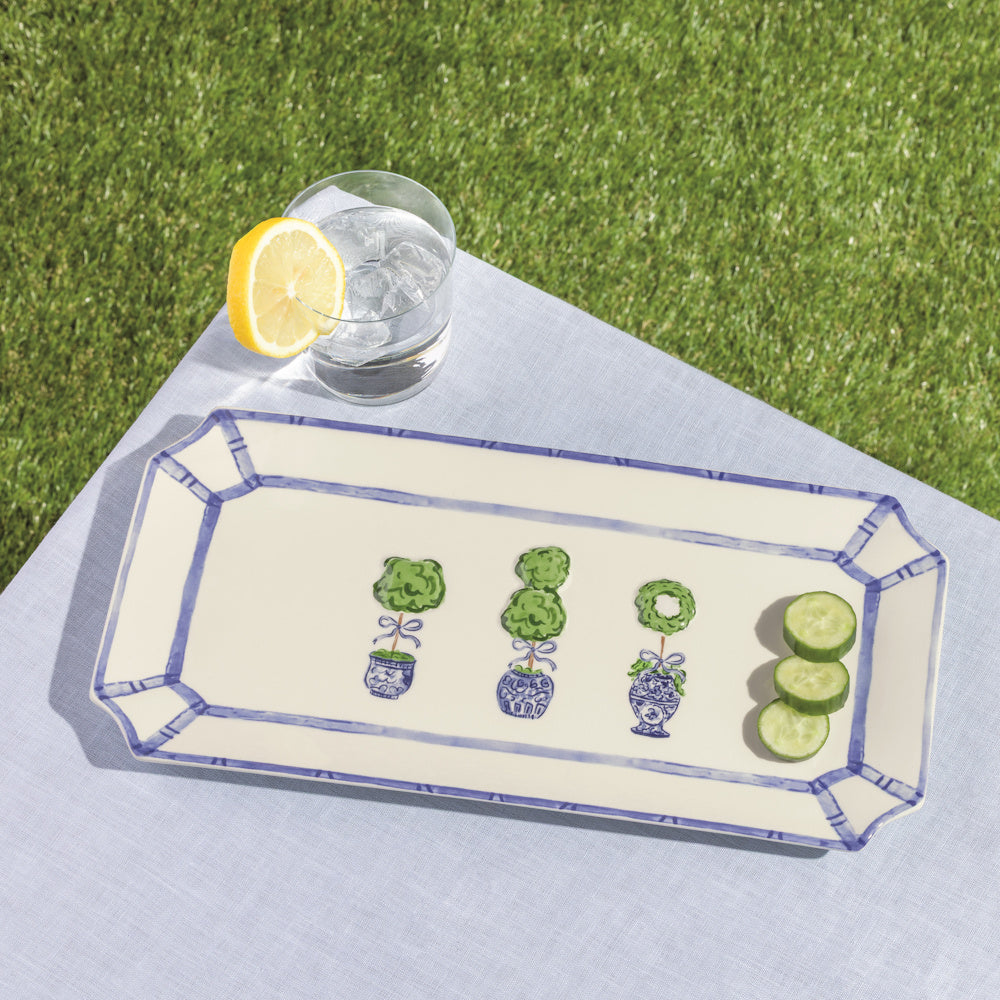 Rectangular ceramic tray with blue bamboo rim and topiary illustrations, featuring cucumber slices and a lemon water glass.
