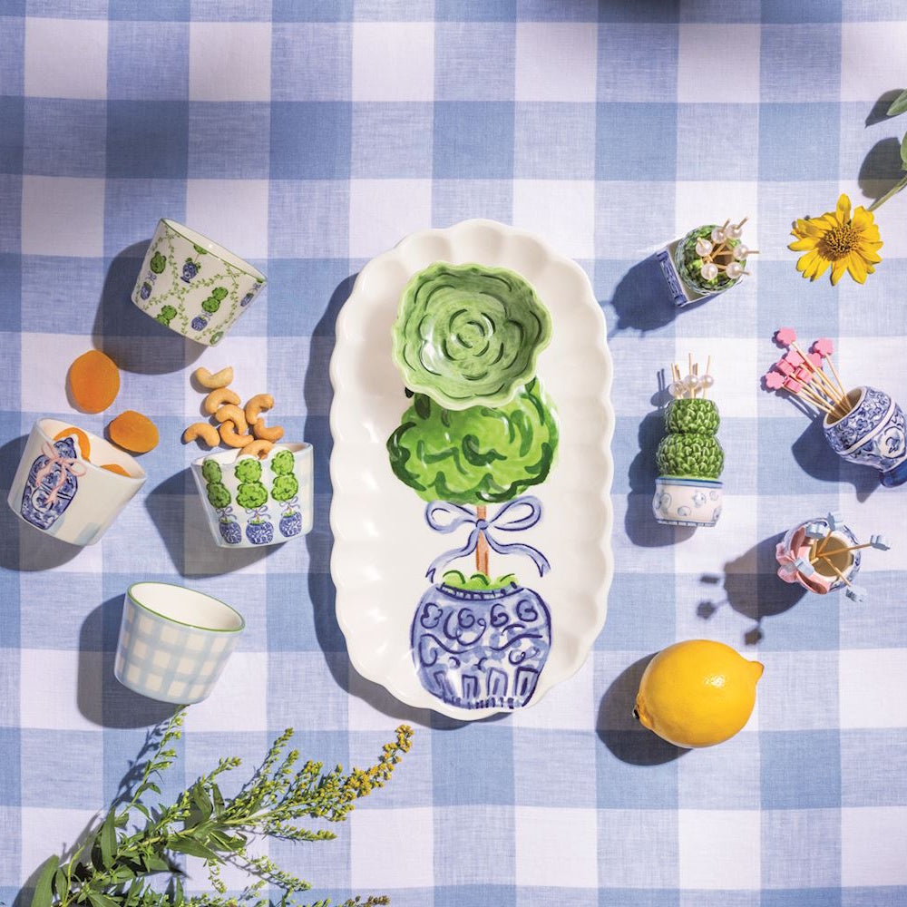 Flat lay of a topiary tidbit tray set with small bowls, snacks, and a lemon on a blue gingham tablecloth.