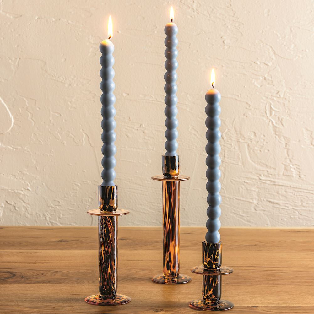 Three tortoise shell glass candlesticks of varying heights with tall blue beaded taper candles on a wooden table.