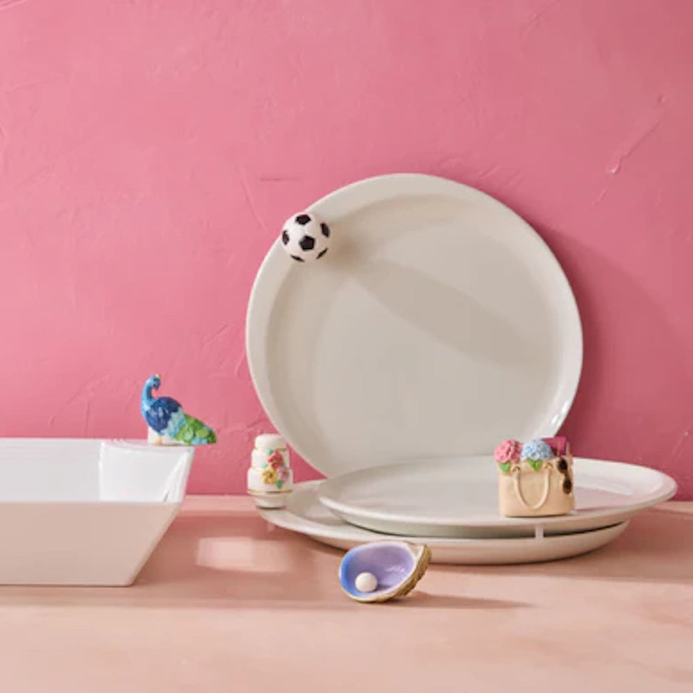 White ceramic serving plates and bowls featuring whimsical decorative charms against a vibrant pink backdrop.