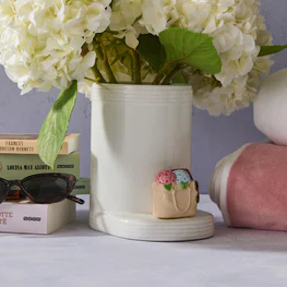 White ceramic vase with hydrangea flowers, a tote bag charm, sunglasses, and books on a table.