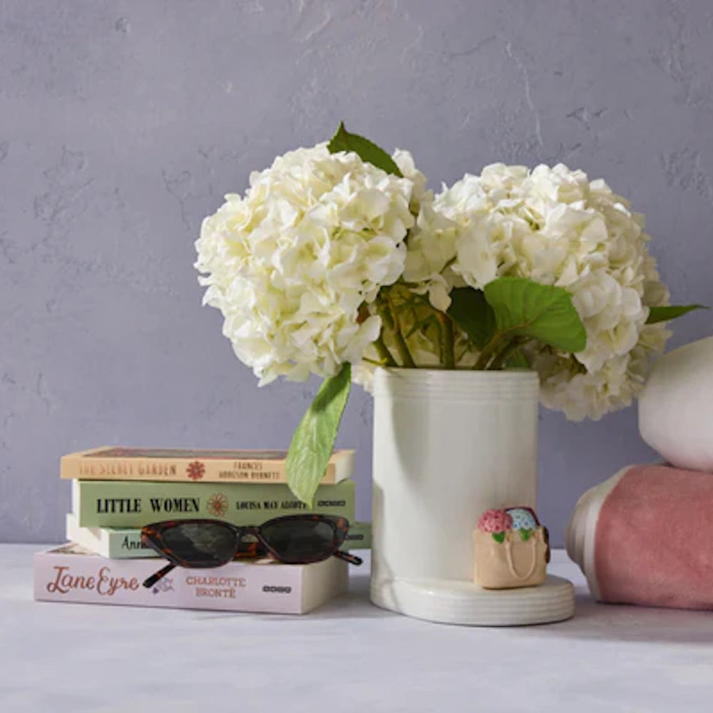 White hydrangeas in a white boot-shaped vase next to a stack of classic books and tortoiseshell sunglasses.