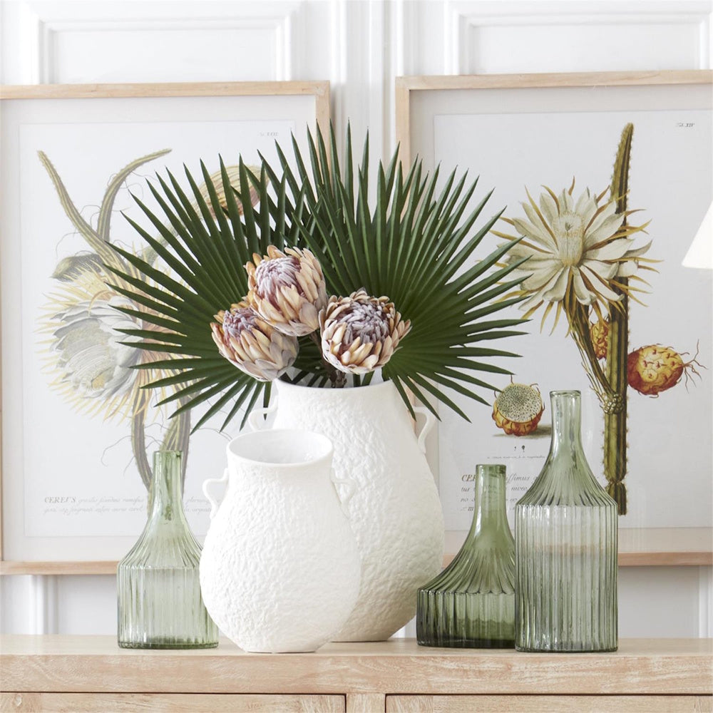 Assorted green ribbed glass and white textured ceramic vases with tropical flowers on a wooden console.
