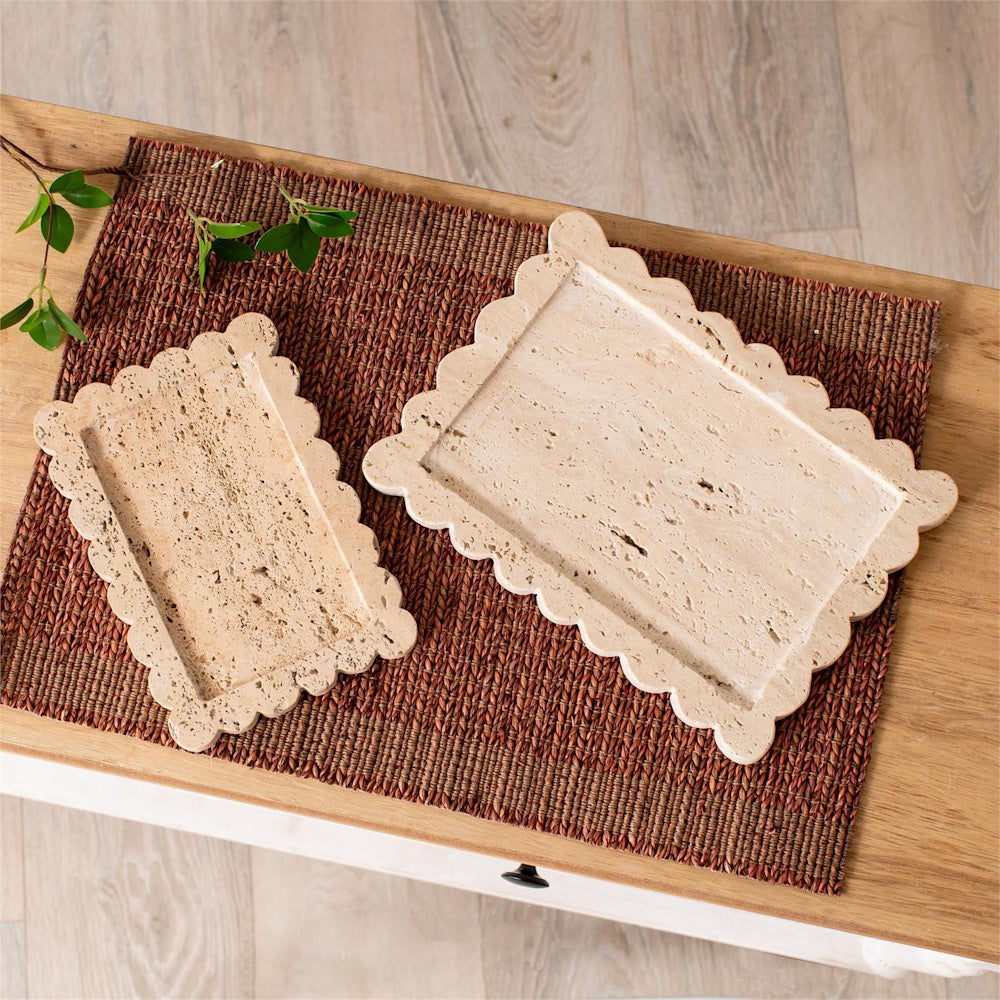 Two scalloped travertine stone decorative trays in different sizes on a textured brown placemat and wooden table.
