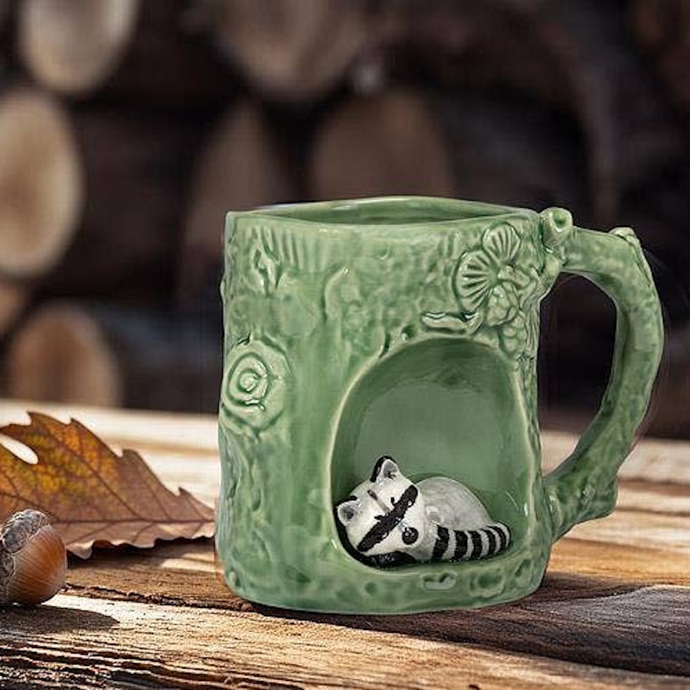 Green ceramic tree stump mug with a sleeping raccoon figurine inside a hollow on a rustic wooden table.