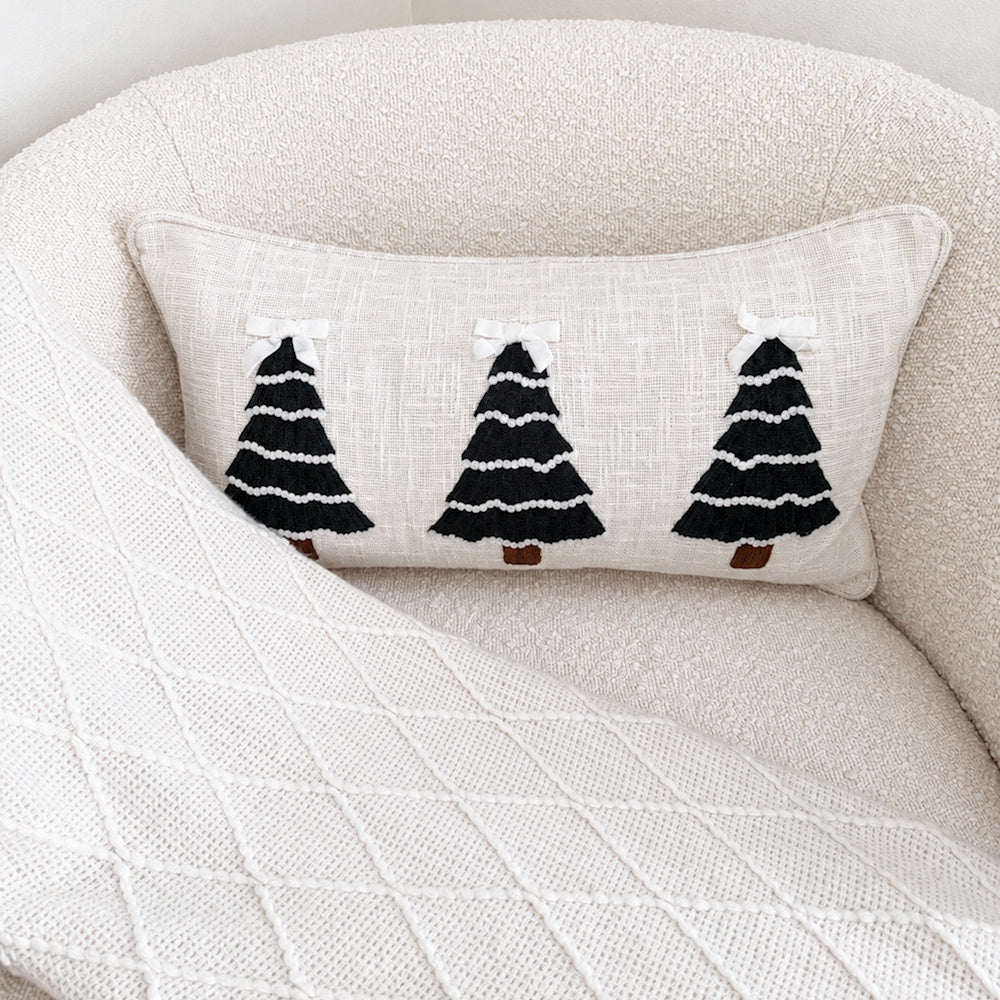Close-up of a Christmas tree embroidered lumbar pillow resting in a white bouclé chair with a throw blanket.