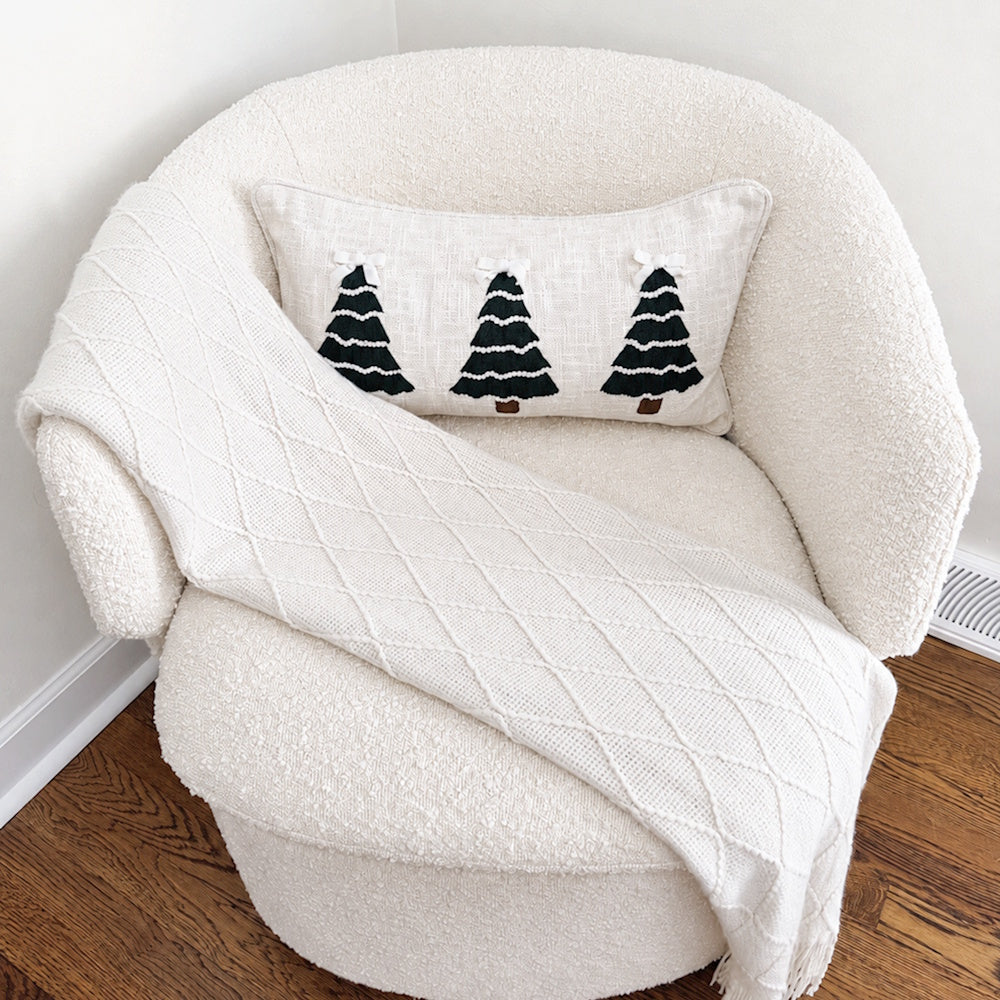 Embroidered Christmas tree pillow styled on a white bouclé armchair with a knit throw blanket.