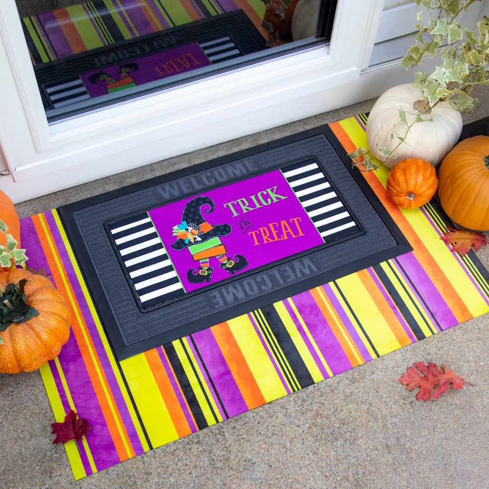Purple witch doormat insert set in a black frame on a colorful striped rug, surrounded by pumpkins.