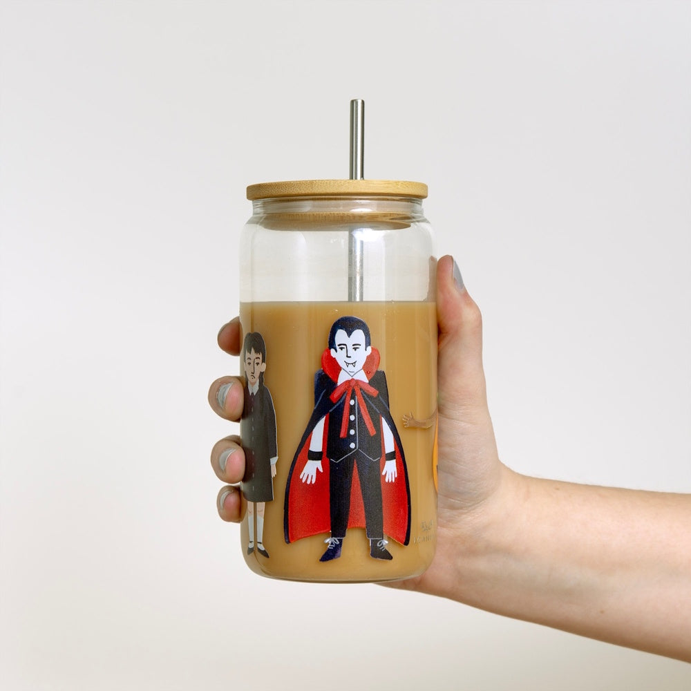 A hand holds a glass can tumbler filled with iced coffee, decorated with Halloween vampire and witch illustrations.