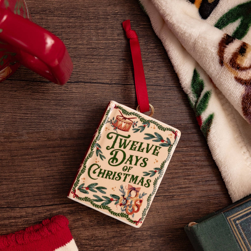 Twelve Days of Christmas ceramic book ornament with a red ribbon on a wooden surface.