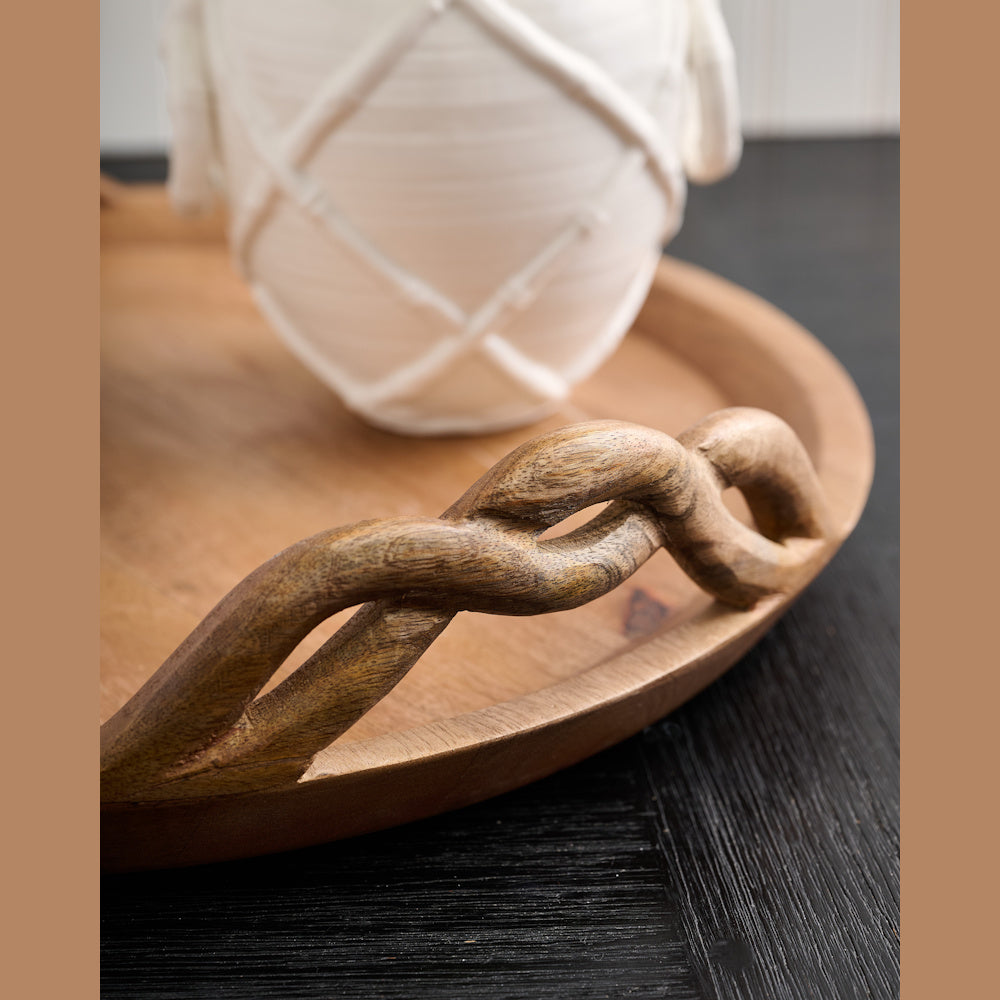 Close-up of a round natural wood tray featuring a carved twisted rope handle on a dark wood table.