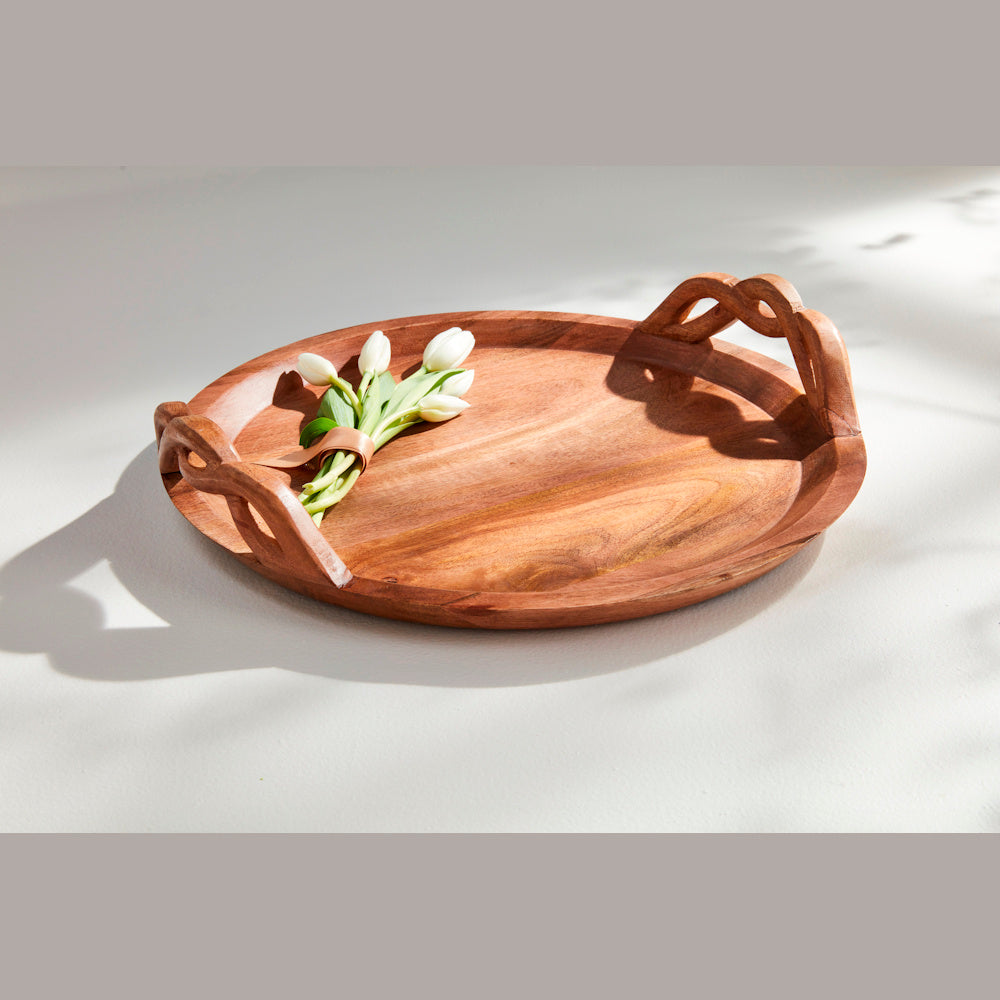 Round acacia wood serving tray with decorative twisted handles and a small bouquet of white tulips.